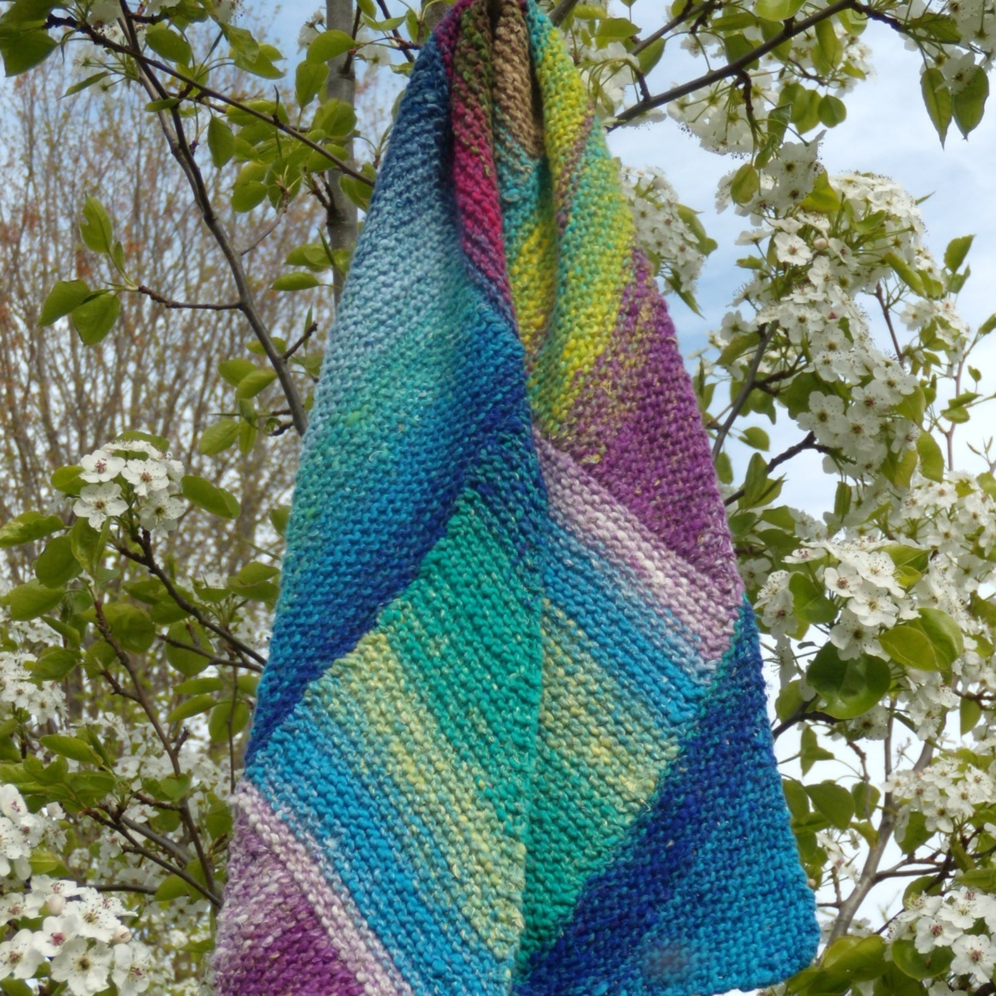 A vibrant Halcyon Yarn Multi-Directional Scarf, downloadable, showcasing a patchwork design in shades of blue, green, purple, and yellow is draped over a tree branch adorned with white blossoms. The background features other branches with green leaves against a partly cloudy sky. This scarf was fun to make and incorporates short rows for added texture.