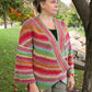 A woman stands outdoors by a tree, wearing the Diagonal Kimono from Halcyon Yarn. The colorful, striped knitted cardigan is made from self-striping worsted weight yarn and features shades of red, orange, green, and pink. She looks to the side with a calm expression, and the background shows a grassy area with trees and fallen leaves.