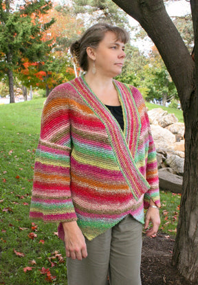 A woman stands outdoors by a tree, wearing the Diagonal Kimono from Halcyon Yarn. The colorful, striped knitted cardigan is made from self-striping worsted weight yarn and features shades of red, orange, green, and pink. She looks to the side with a calm expression, and the background shows a grassy area with trees and fallen leaves.