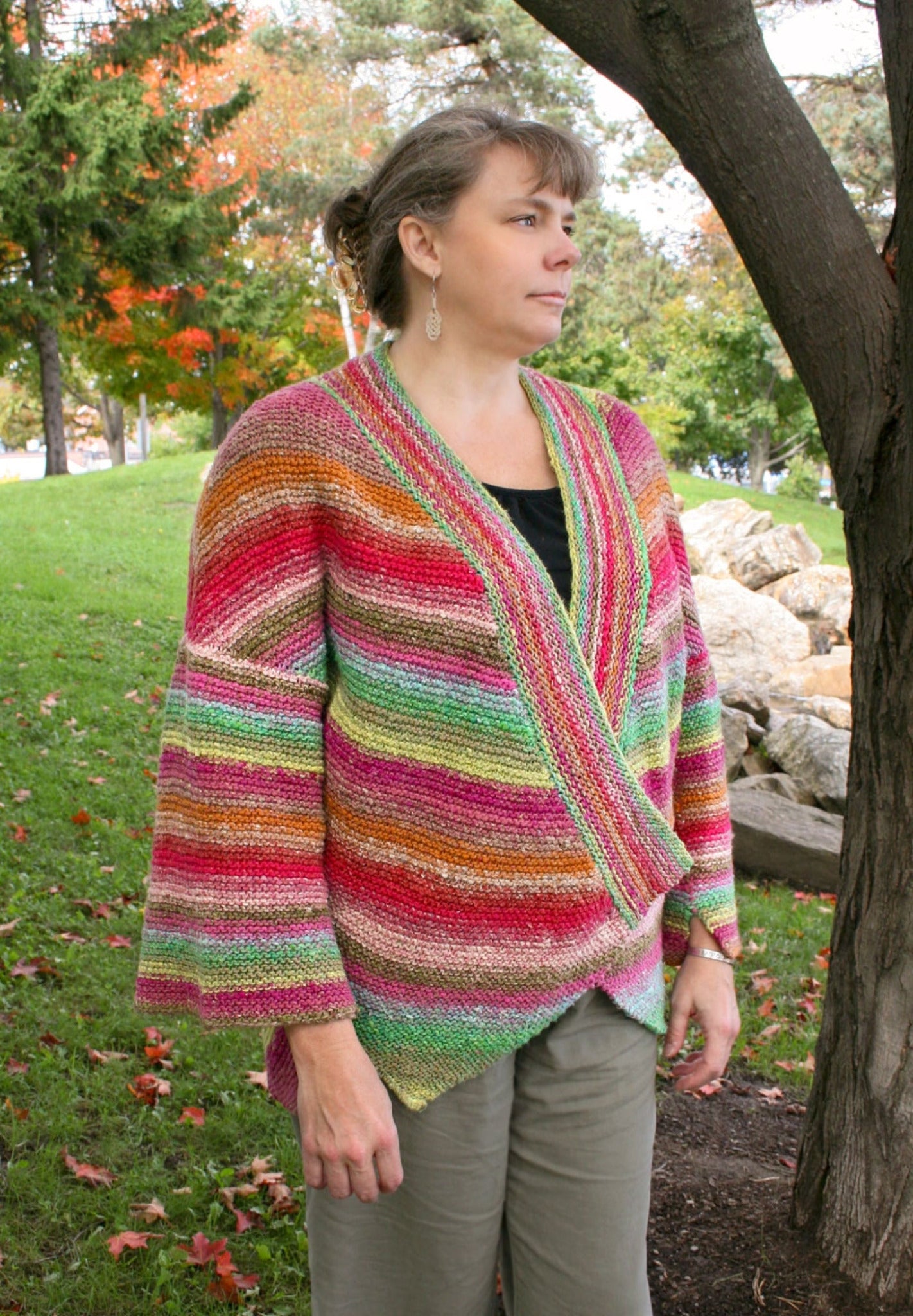 A person is standing outdoors near a tree, wearing the Diagonal Kimono by Halcyon Yarn. The colorful, striped knit cardigan is crafted from self-striping worsted weight yarn in green, pink, red, and beige hues. They have their hair tied back and are looking to the side. The background includes a grassy area with scattered leaves and more trees.