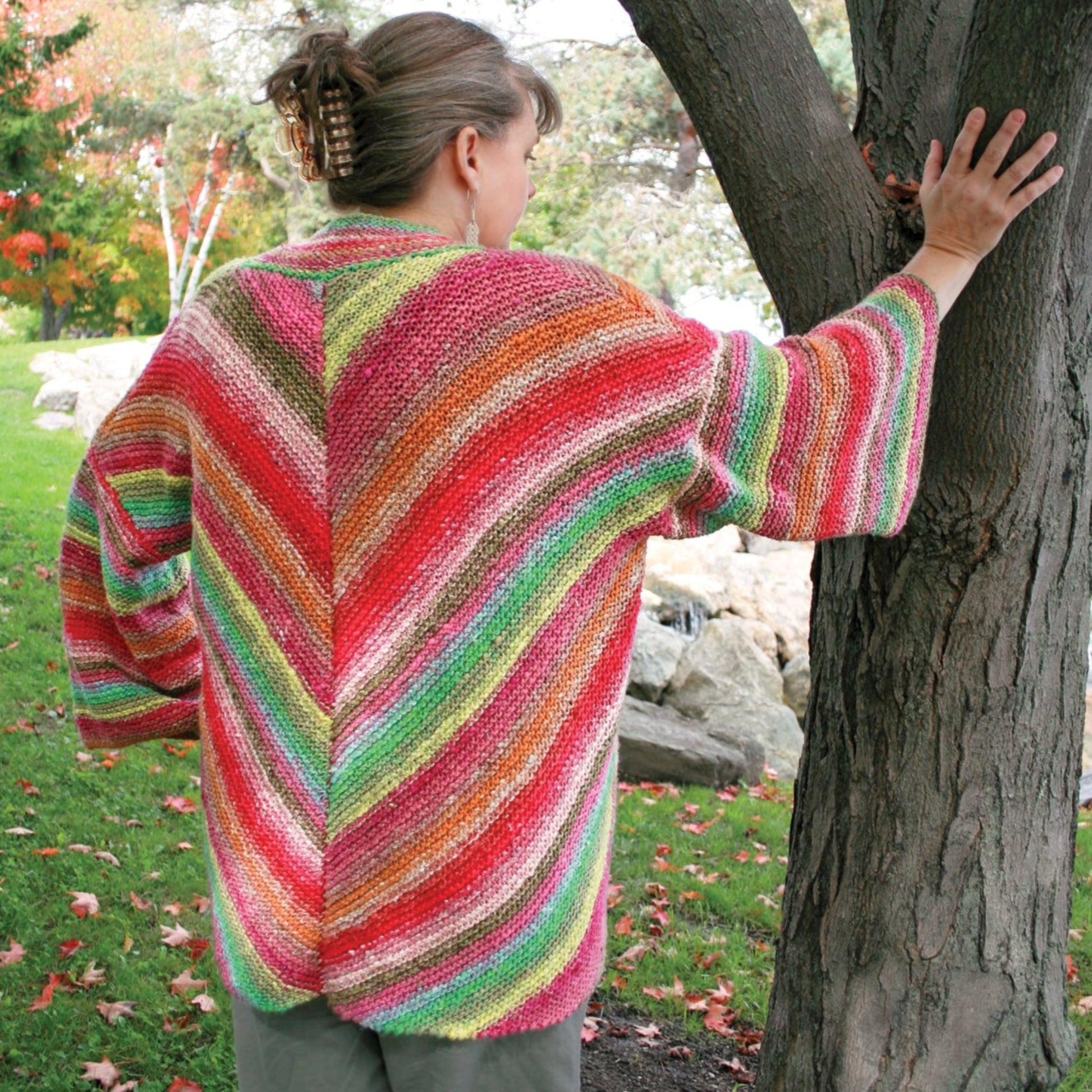 A person with short hair pinned up is standing outdoors near a tree, facing away from the camera. They are wearing the Halcyon Yarn Diagonal Kimono, a multicolored, striped sweater made from self-striping yarn with wide sleeves. The background shows a grassy area with some fallen leaves and other trees.