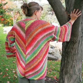 A person stands outdoors with their back to the camera, donning the Diagonal Kimono from Halcyon Yarn, a vibrant, self-striping worsted weight yarn cardigan featuring shades of red, green, yellow, and pink. They rest one hand on a tree trunk while their hair is styled up with a tortoiseshell hair clip.