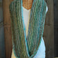 A Halcyon Yarn Transitions Cowl, featuring green, blue, and white horizontal stripes, is displayed on a wicker mannequin. The mannequin is also dressed in a plain white T-shirt and positioned against a backdrop of rustic wooden planks.