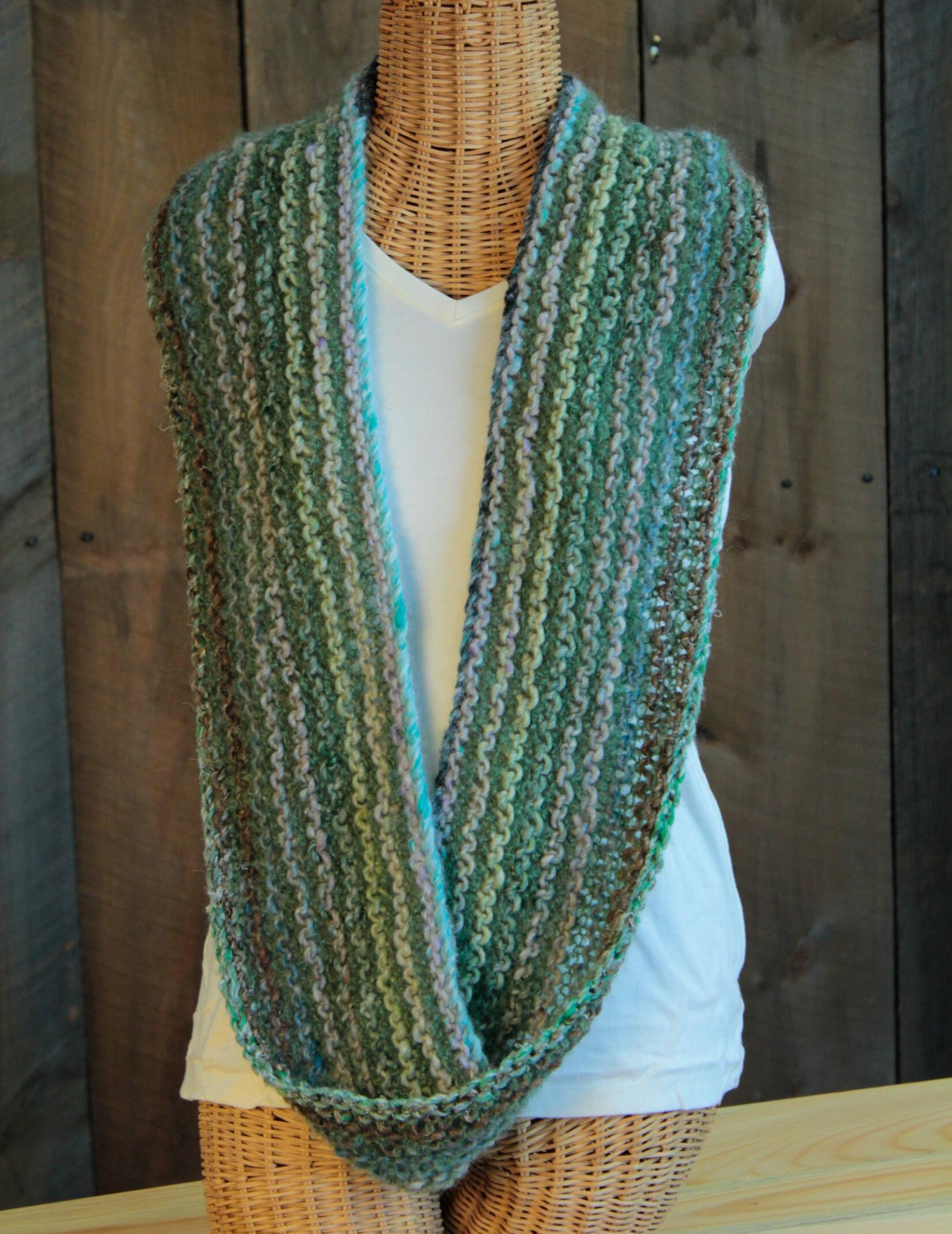 A Halcyon Yarn Transitions Cowl, featuring green, blue, and white horizontal stripes, is displayed on a wicker mannequin. The mannequin is also dressed in a plain white T-shirt and positioned against a backdrop of rustic wooden planks.