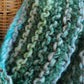 Close-up of a basket and the Transitions Cowl, hand-knit with a textured, yarn pattern in earthy green, teal, and brown tones. The cozy Victorian Boucle knit by Halcyon Yarn showcases a variety of green hues and appears soft and warm.