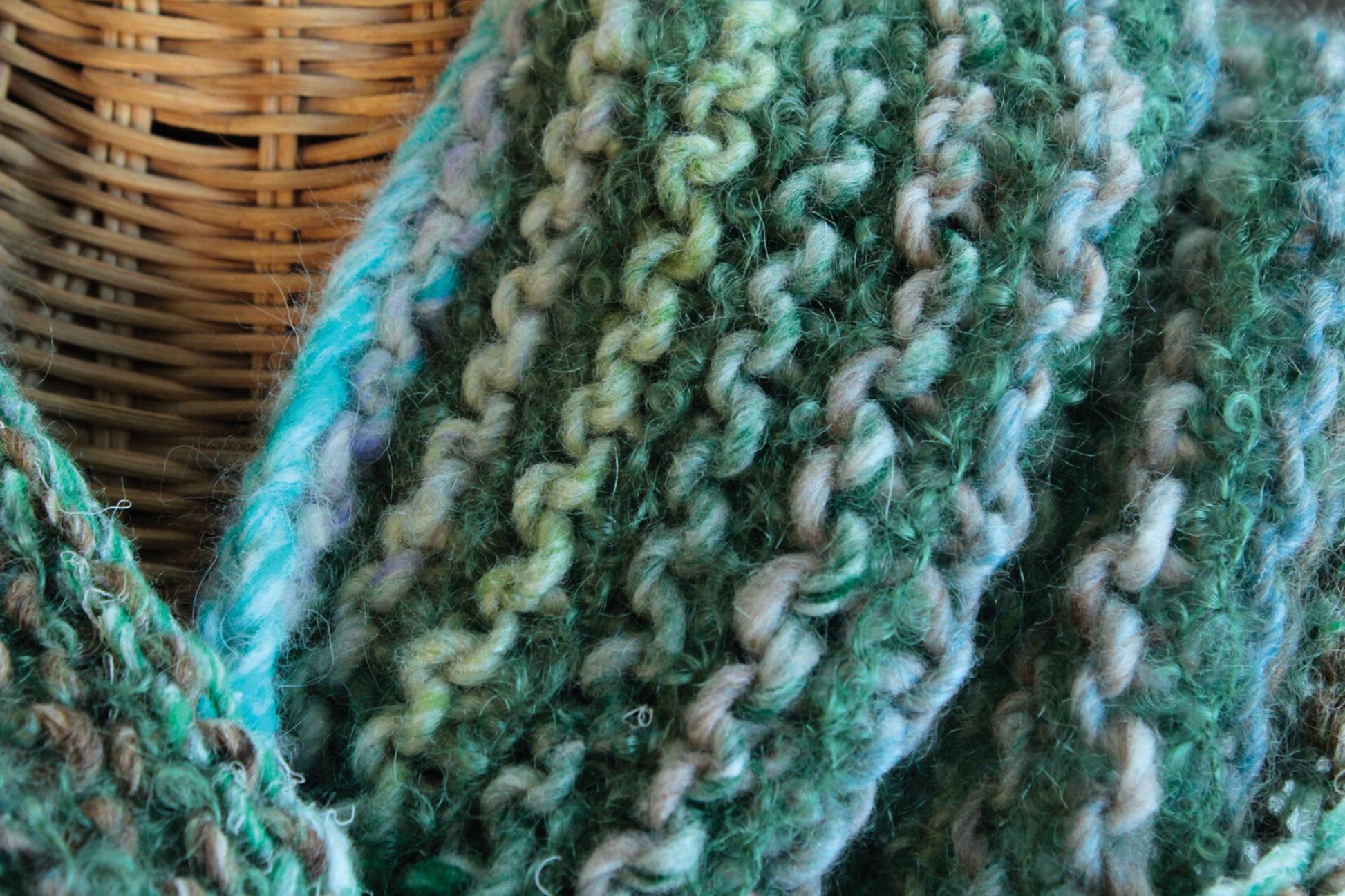 Close-up of a basket and the Transitions Cowl, hand-knit with a textured, yarn pattern in earthy green, teal, and brown tones. The cozy Victorian Boucle knit by Halcyon Yarn showcases a variety of green hues and appears soft and warm.