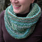 A person with a partial smile is wearing a hand-knitted Transitions Cowl from Halcyon Yarn. The multi-colored green and brown cowl, which appears cozy and textured, wraps loosely around their neck over a dark top, showcasing an array of blended colors and intricate knitting patterns.