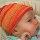 A baby wearing Halcyon Yarn's "Baby's First Hat" in an orange and red striped pattern lies down, gazing to the right. Dressed in a light-colored outfit, the baby has their hand near their face, against a soft, off-white blanket background.