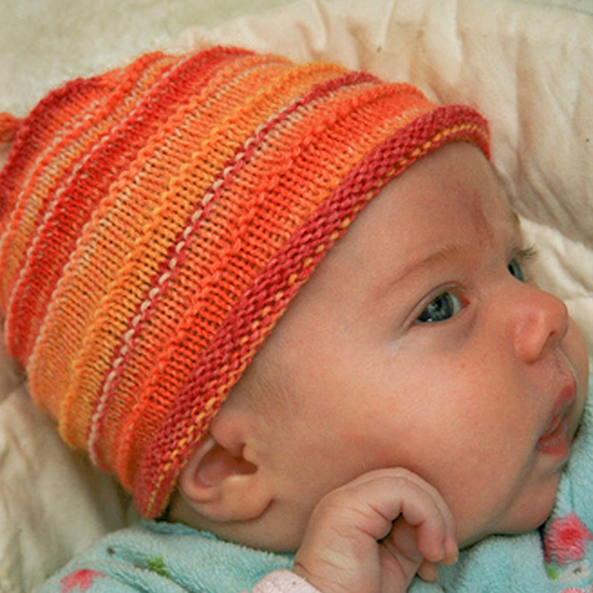 A baby wearing Halcyon Yarn's "Baby's First Hat" in an orange and red striped pattern lies down, gazing to the right. Dressed in a light-colored outfit, the baby has their hand near their face, against a soft, off-white blanket background.