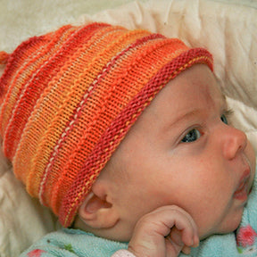 Close-up of a baby wearing Halcyon Yarn's Baby's First Hat, featuring a handknit design in vibrant self-striping orange and red hues, as they look to the side. The baby’s subtle expression is accentuated by their resting hand near their chin against a soft and cushioned background.