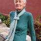 An older woman with short, white hair, wearing a teal turtleneck sweater and a Halcyon Yarn Cashmere Confection Scarf, sits on a stone bench. She poses with a thoughtful expression with a red wall and some greenery visible in the background.