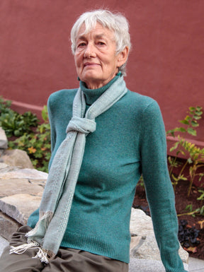 An older woman with short, white hair, wearing a teal turtleneck sweater and a Halcyon Yarn Cashmere Confection Scarf, sits on a stone bench. She poses with a thoughtful expression with a red wall and some greenery visible in the background.