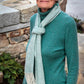 An elderly woman with short gray hair, wearing a teal Jade Sapphire Cashmere sweater and a Cashmere Confection Scarf by Halcyon Yarn, stands outdoors in front of a stone wall and some greenery. She has an expression of quiet reflection.