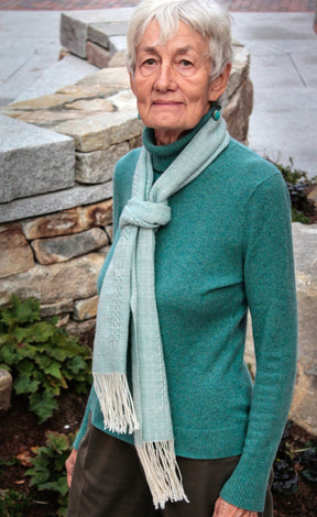 An elderly woman with short gray hair, wearing a teal Jade Sapphire Cashmere sweater and a Cashmere Confection Scarf by Halcyon Yarn, stands outdoors in front of a stone wall and some greenery. She has an expression of quiet reflection.