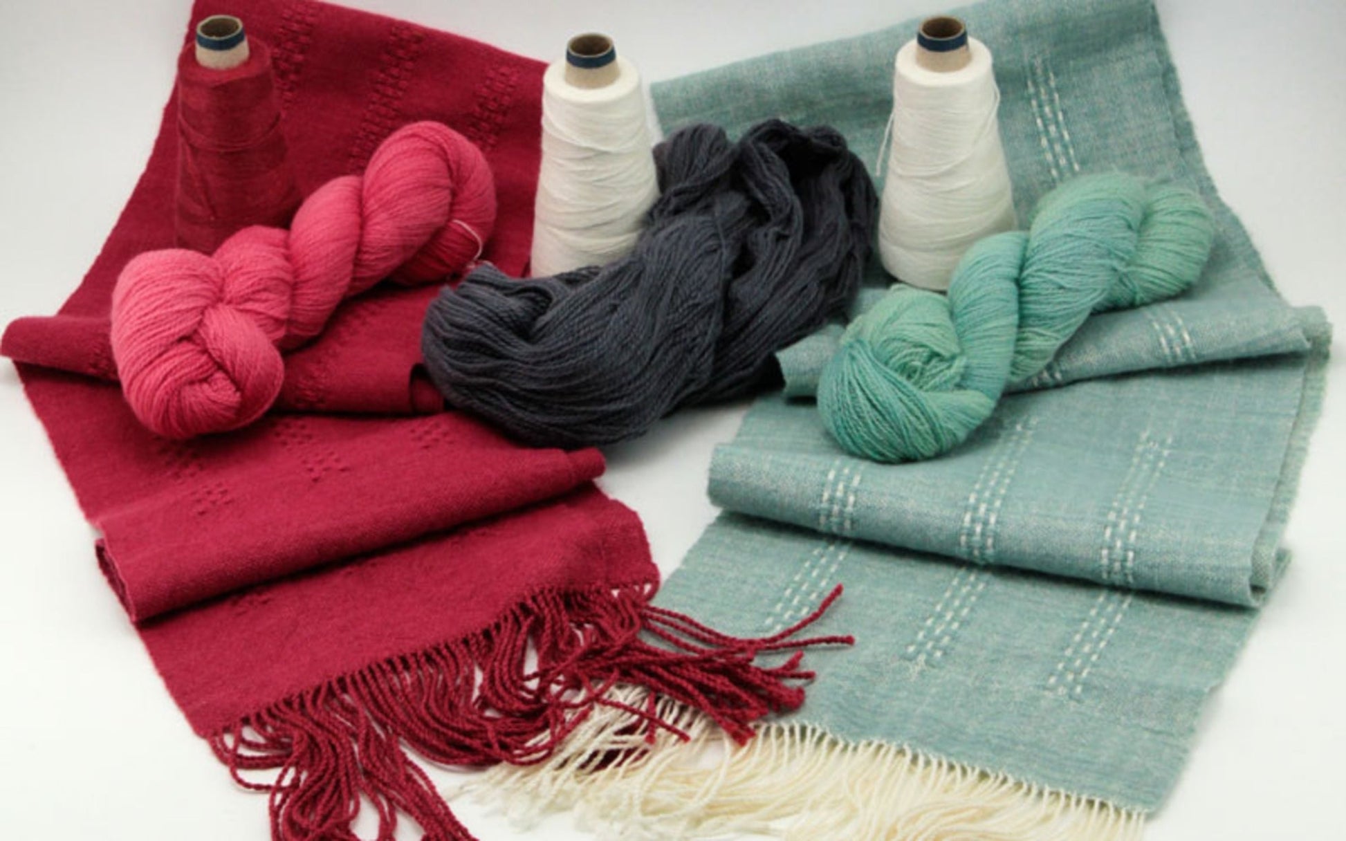 A display of Halcyon Yarn's Cashmere Confection Scarf in red and teal woven textiles features fringe ends on the red version and light stripes on the teal. Complementing the scarves are matching Jade Sapphire Cashmere yarn skeins and Zephyr wool/silk thread spools.