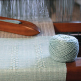 A ball of light green yarn sits on a four-shaft loom with partially woven fabric of the same color, destined to become the "Cashmere Confection Scarf" by Halcyon Yarn. The loom has wooden parts, and the warp threads are stretched taut. The yarn is neatly wound, and the woven fabric shows a simple, elegant texture pattern.