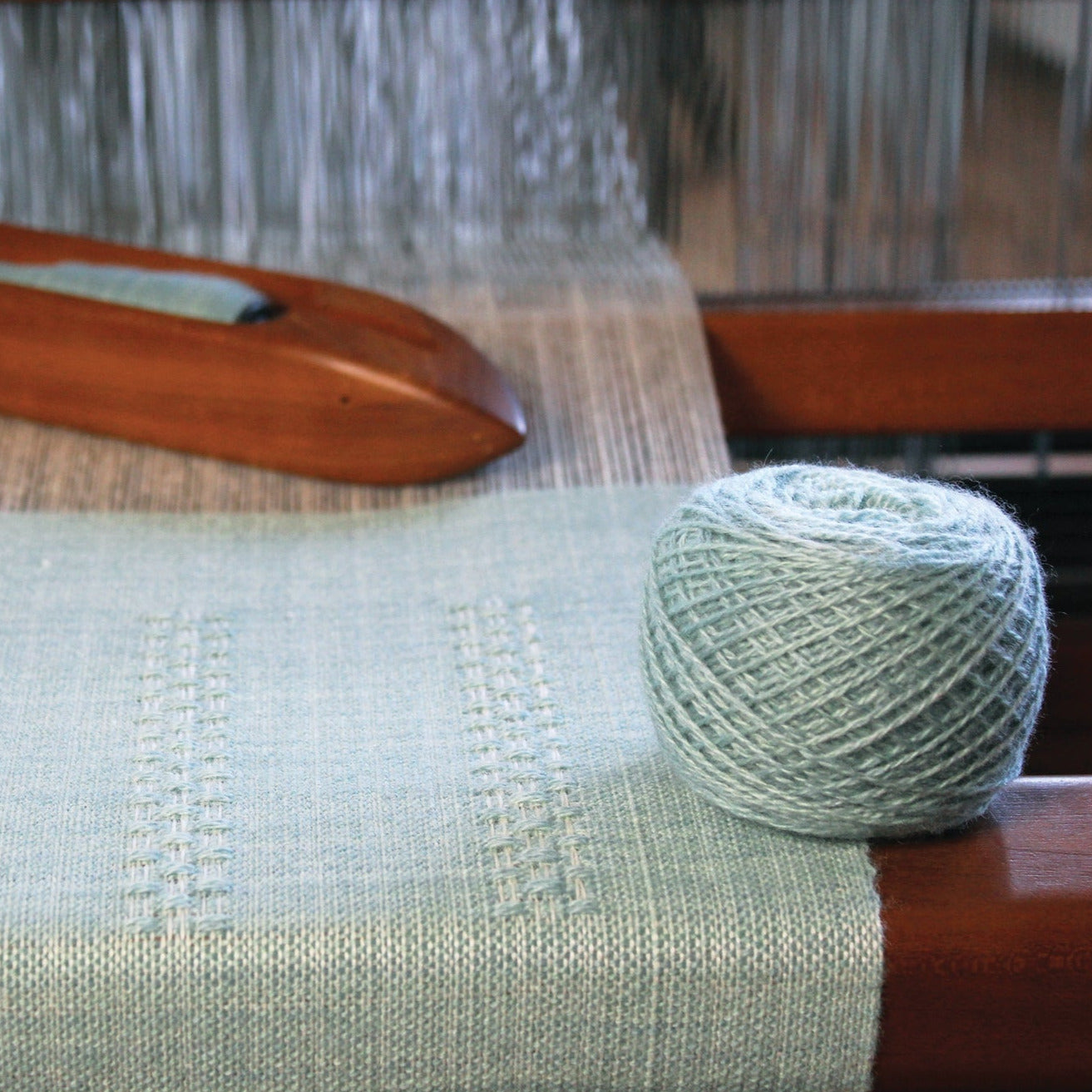 A ball of light blue Zephyr wool/silk yarn rests on a wooden loom, where a piece of light blue woven fabric is in progress. The loom shuttle peeks out in the background, partially woven into what promises to be a beautiful huck lace scarf from the Cashmere Confection Scarf pattern by Halcyon Yarn, showcasing the intricate weaving process.