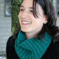 A person with shoulder-length dark hair and a smile is wearing the Halcyon Yarn Cashmere Cowl with a Twist in teal laceweight and stud earrings. They are dressed in a black top and are standing outdoors in front of a tree trunk and a building.