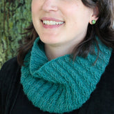 A person is wearing the Cashmere Cowl with a Twist from Halcyon Yarn in green. Paired with a black top, they are smiling, with only the lower part of their face visible. The background features a blurry tree trunk. This lace-weight cashmere scarf boasts a ribbed pattern, adding an extra touch of cozy elegance.