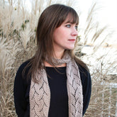 A woman with straight brown hair and bangs stands in front of tall, dry grass, looking to the left. She wears a black top and a light brown, luxurious Shell Lace Scarf by Halcyon Yarn, appearing to be in an outdoor setting with a calm, cloudy sky.