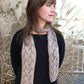 A person with shoulder-length brown hair and bangs stands outdoors, looking to the left. They are wearing a black long-sleeve shirt and a luxurious Shell Lace Scarf in light grey from Halcyon Yarn. Behind them are tall, dry grass and a blurred background.