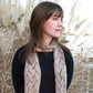 A woman with shoulder-length brown hair and green eyes wearing a black long-sleeve top and the luxurious Shell Lace Scarf from Halcyon Yarn stands in front of tall, dried grass. She is looking to her left with a thoughtful expression.