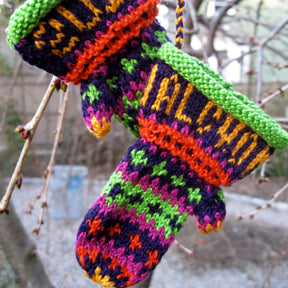 Halcyon Yarn's Christmas Mitten Ornament, with its intricate patterns in vibrant shades of purple, green, red, orange, and yellow, complemented by green cuffs, hangs colorfully on a bare tree branch. The background showcases blurred branches and a garden area.