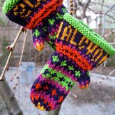 Two vibrant Christmas Mitten Ornaments from Halcyon Yarn dangle from a tree branch, featuring festive patterns with stripes and stars in green, purple, orange, and red. Crafted from sport weight yarn, these colorful decorations contrast beautifully with the stark branches around them.