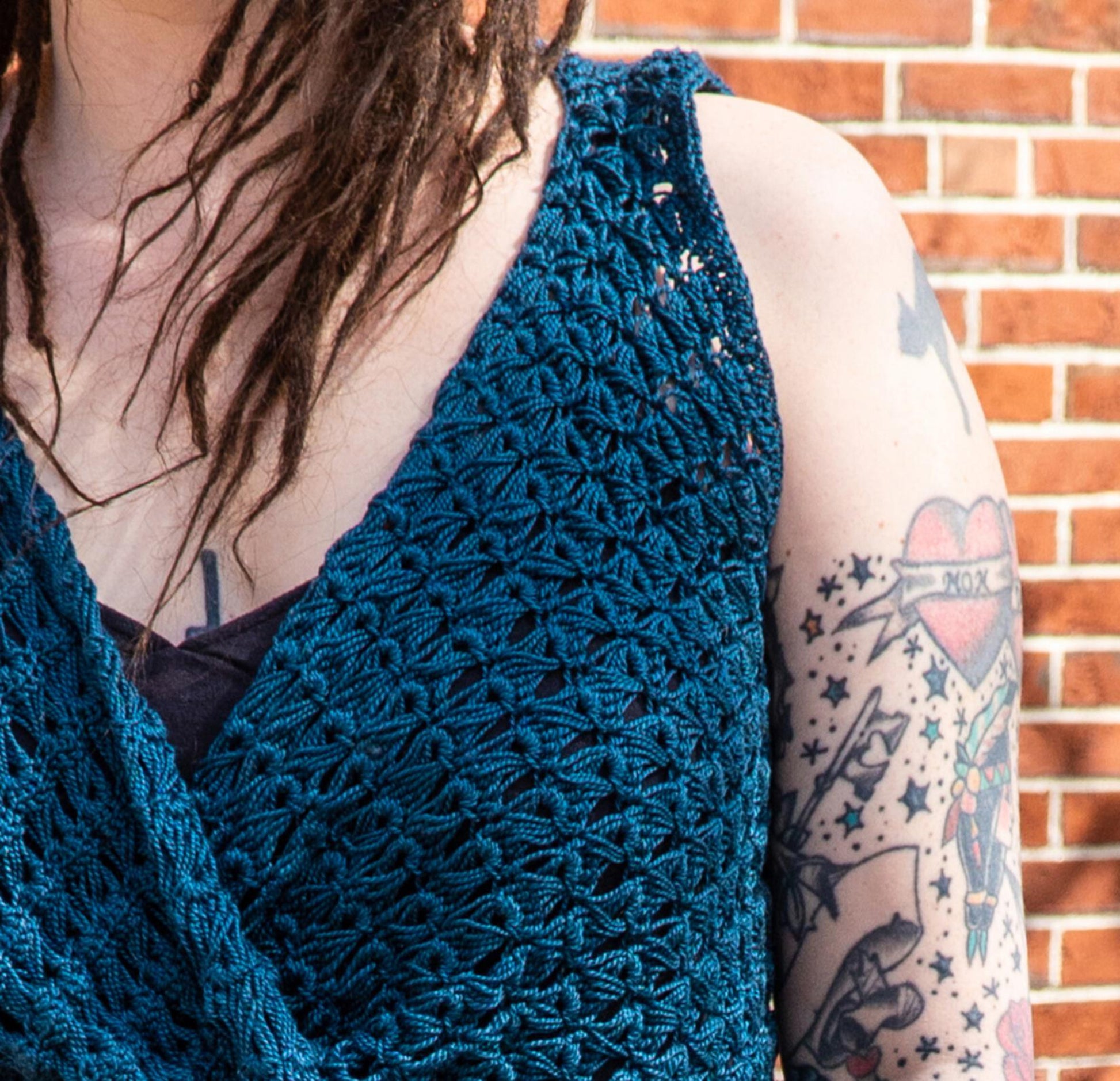 Close-up of a person's upper torso and arm. They are wearing the Ultra Twist Broomstick Lace Crochet Top by Halcyon Yarn, crafted from sport weight yarn in a striking blue. Their arm is adorned with multiple tattoos, including a heart with a banner and smaller designs. The background is a rustic brick wall.