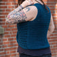 A person with tattoos on their arms is facing away from the camera, showing off the Ultra Twist Broomstick Lace Crochet Top from Halcyon Yarn in dark teal. They are touching their hair, and there is a brick wall in the background.