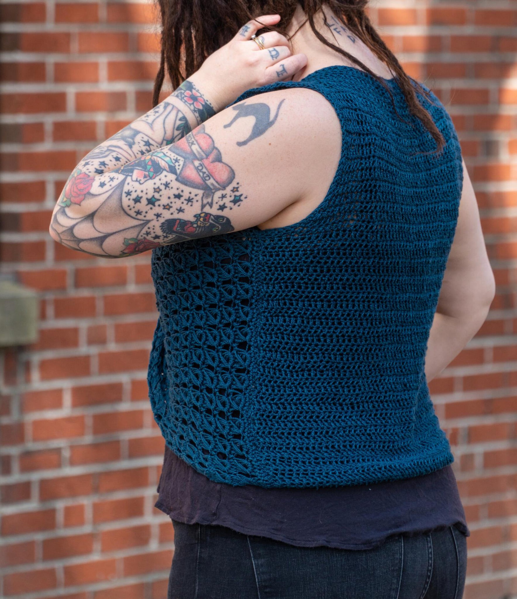 A person with tattoos on their arms and hand stands facing away from the camera, wearing an Ultra Twist Broomstick Lace Crochet Top in a deep blue color made from sport weight yarn by Halcyon Yarn. They are touching their hair with their left hand, with a brick wall visible in the background.