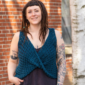 A smiling person stands outside in front of a brick wall and a tree, wearing Halcyon Yarn's Ultra Twist Broomstick Lace Crochet Top: a dark tank under a blue double crochet vest. They have long dreadlocks adorned with beads and show multiple tattoos on both arms.