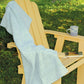 A light wooden Adirondack chair is positioned outdoors on a grassy area. A soft blue Woven Bath Sheet from Halcyon Yarn is draped over the back and seat of the chair. A blue and white ceramic mug and an open book rest on one of the chair's armrests.