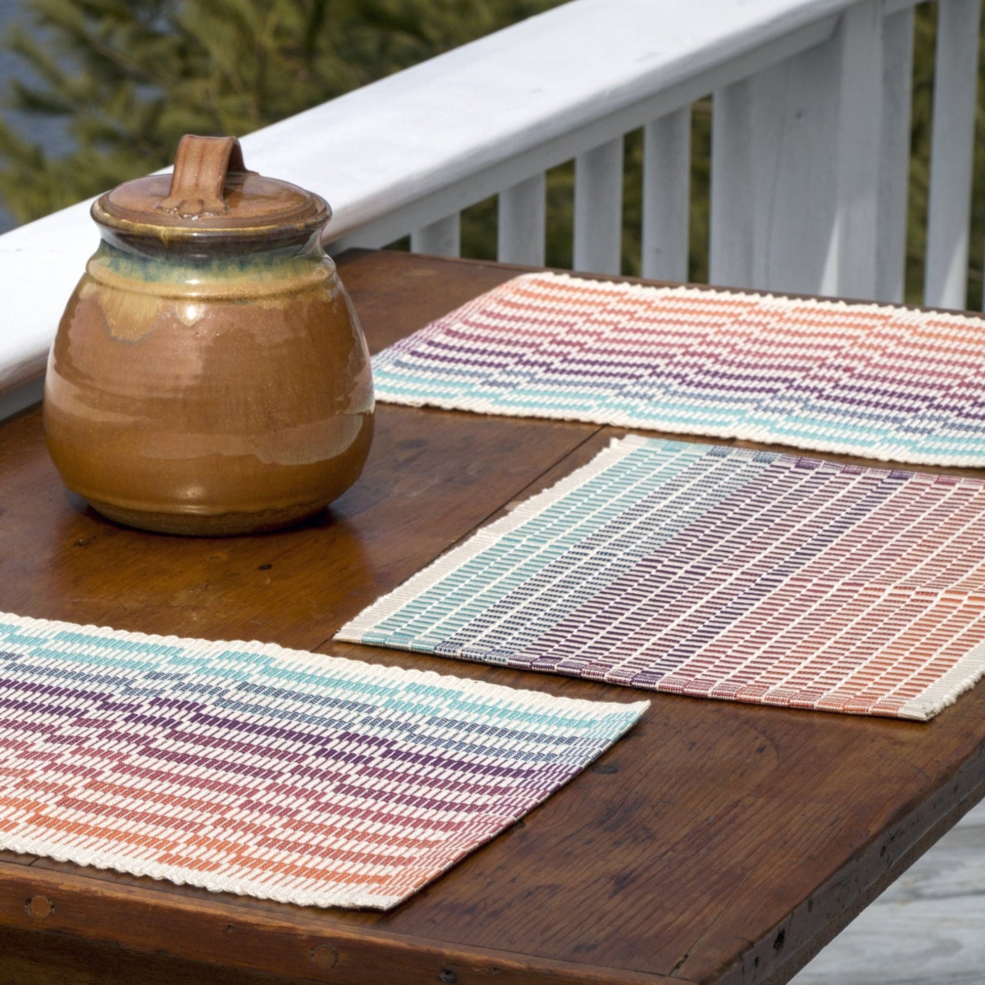 A wooden table on an outdoor porch is adorned with three Lovely Day Rep Weave Placemats from Halcyon Yarn, each featuring gradient patterns in shades of purple, blue, green, and orange. A ceramic pot with a lid sits on the table. In the background, a white railing and green foliage are visible.