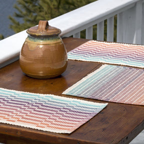 A wooden table on a balcony features three vibrant Lovely Day Rep Weave Placemats from Halcyon Yarn, crafted on a 4-shaft loom, alongside a ceramic jar with a lid. A white railing and lush green foliage can be seen in the background, creating a peaceful outdoor setting.