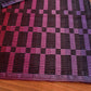 A close-up of the Rep Weave Placemat Pattern by Halcyon Yarn showcases a geometric design with horizontal and vertical stripes in shades of black, purple, and pink. The textured surface of the Rep Weave structure makes it an ideal choice for placemats and runners. A wooden surface is partially visible in the bottom right corner.