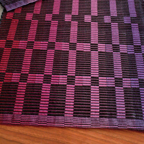 A close-up of the Rep Weave Placemat Pattern by Halcyon Yarn showcases its geometric design. This fabric is perfect for crafting placemats and runners, featuring an array of vertical and horizontal stripes in diverse shades of purple, pink, and black. The ribbed texture creates a stunning surface, with the edges clearly visible against a wooden backdrop.