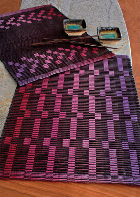 A dining table is set with two rectangular Halcyon Yarn Rep Weave Placemats featuring a gradient pattern of purples and reds in a geometric design. On the table are also two square dishes with a gradient from turquoise to brown, and a pair of chopsticks resting on one.