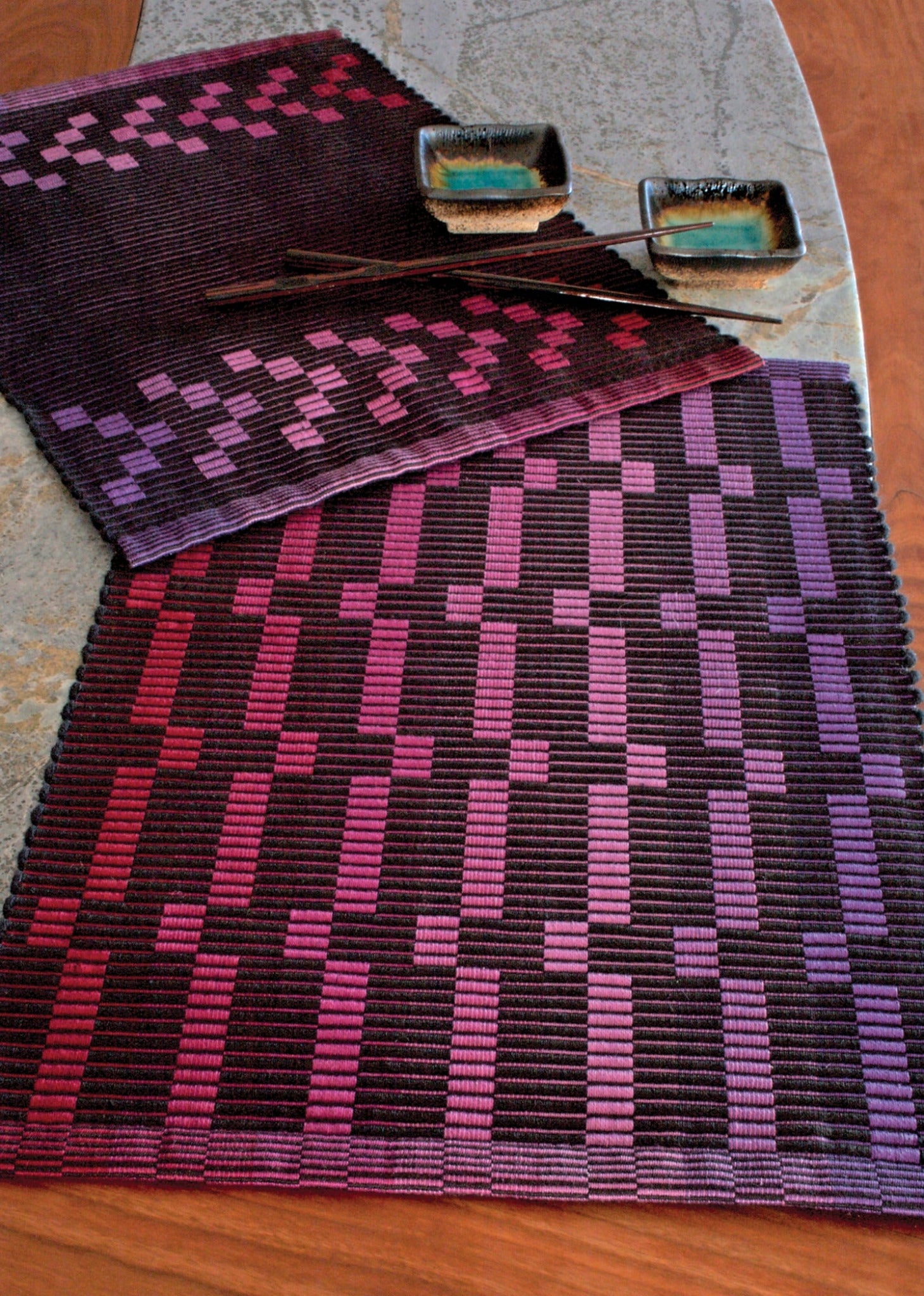 Three woven Rep Weave Placemat Pattern pieces from Halcyon Yarn, featuring a geometric design in various shades of red, purple, and pink, are displayed on a beautiful surface. Two dishes with colorful glazes rest on the placemats and runners, adding a touch of contrast and highlighting the artistic texture.