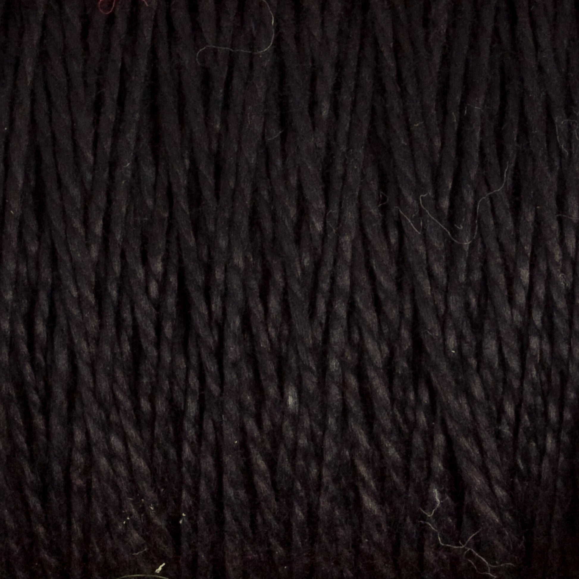 Close-up view of tightly wound, dark-colored Supreme Corp.'s 10/2 Pearl Cotton Yarn on a large cone. The strands are uniformly thick and tightly packed together, showcasing a consistent texture across the image with slight variations in shading due to lighting. This versatile yarn, often used in pearl cotton projects, offers durability and rich hues.