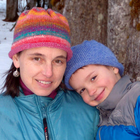 A woman and a young boy stand close together outdoors, both sporting colorful Classic Roll Brim Hats from Halcyon Yarn and warm jackets. The boy smiles and leans his head affectionately against the woman's shoulder as they enjoy a snowy, wooded area.