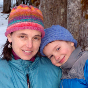 A woman and a child, both wearing Halcyon Yarn Classic Roll Brim Hats and warm jackets, smile at the camera while standing outside in a snowy, wooded area. The woman sports a colorful hat, while the child wears a blue one suitable for all ages, leaning affectionately against the woman.