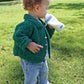 A young child with curly hair stands on grass, wearing a green knitted Ellen's Favorite Baby Sweater by Halcyon Yarn made from Casco Bay Chenille Worsted, blue jeans, and white shoes with a flower pattern. The child is holding a baby bottle and drinking from it while looking down. Trees and buildings are visible in the background.