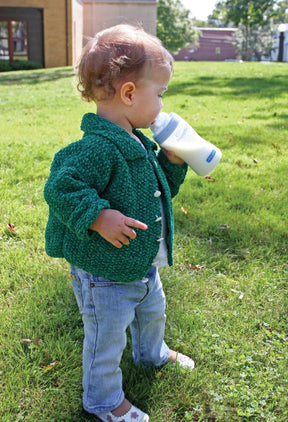 A young child with curly hair stands on grass, wearing a green knitted Ellen's Favorite Baby Sweater by Halcyon Yarn made from Casco Bay Chenille Worsted, blue jeans, and white shoes with a flower pattern. The child is holding a baby bottle and drinking from it while looking down. Trees and buildings are visible in the background.