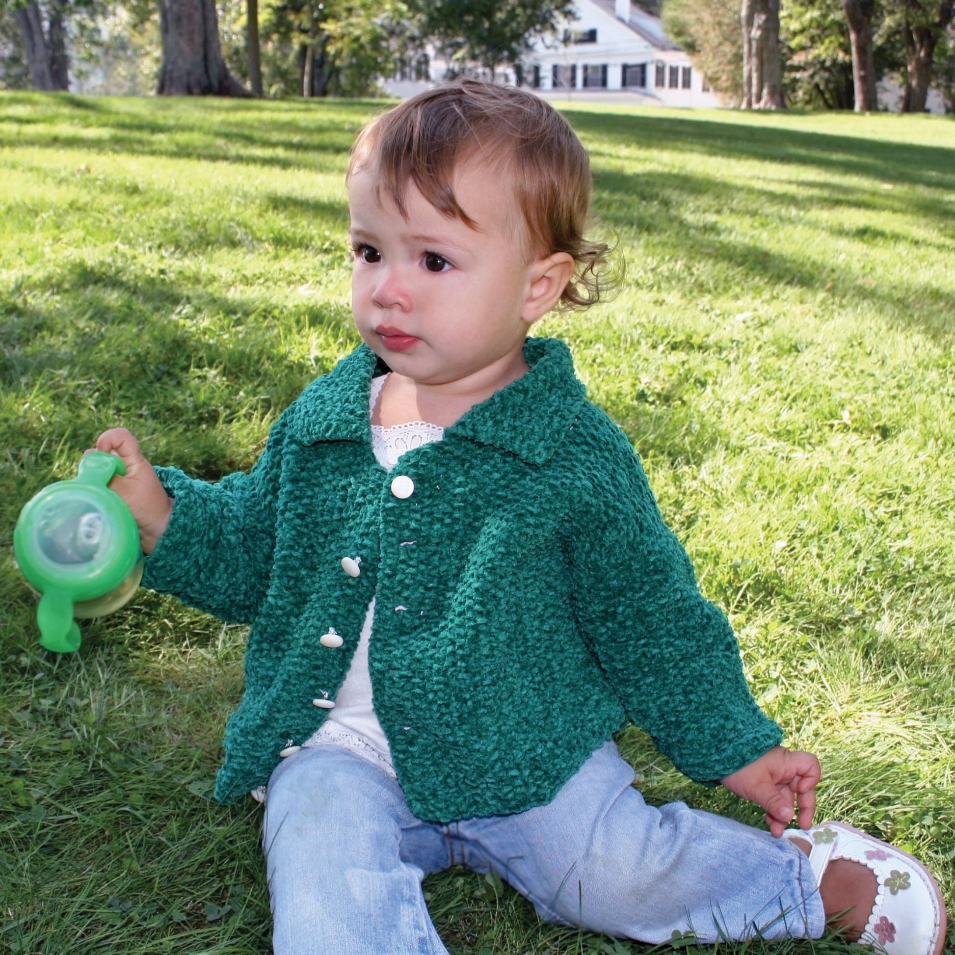 A toddler with short hair, wearing Ellen's Favorite Baby Sweater by Halcyon Yarn made from Casco Bay Chenille Worsted, light blue jeans, and white shoes, sits on the grass in a park holding a green sippy cup. Trees and a house are visible in the background.