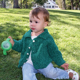 A young child sits on green grass, holding a green sippy cup with their right hand. The child is wearing Halcyon Yarn's Ellen's Favorite Baby Sweater, along with light blue jeans and white shoes. In the background, tall trees sway near a white house with dark shutters.