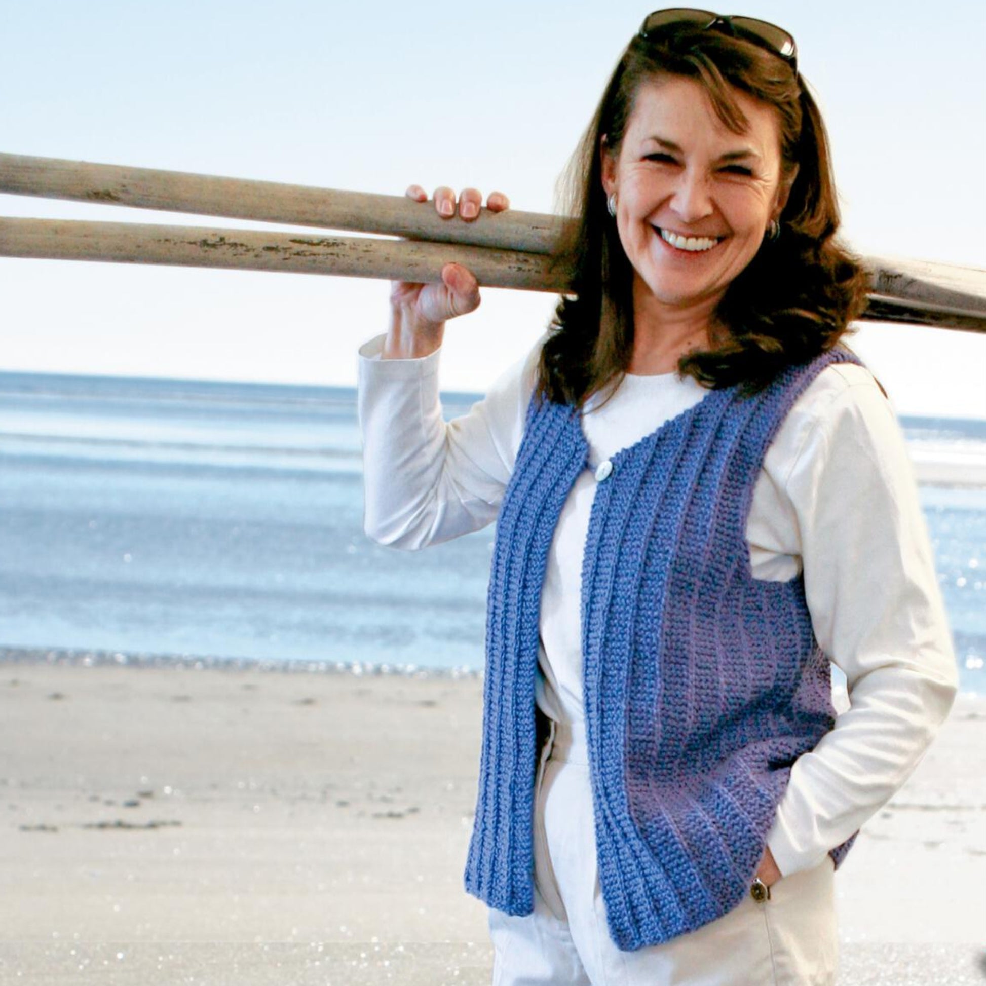 A woman with long brown hair and sunglasses on her head smiles while holding two oars over her shoulder, dressed in a white shirt and the Halcyon Yarn Vaill Island Vest, downloadable in a garter slip stitch pattern. The background reveals a sandy beach with a calm blue ocean and a clear sky.