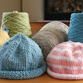 A collection of Casco Bay Baby Roll Brim Hats from Halcyon Yarn is displayed on a table, featuring a soft cotton baby hat in blue and a pink-and-white striped hat. Behind them are large spools of Casco Bay Sport yarn in colors like green, beige, and blue. The cozy indoor setting is enhanced with free knitting patterns scattered around.