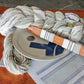 A coiled rope and a wooden weaving shuttle lie on top of an intricately crafted Rustic Mats, Runner & Napkins from Halcyon Yarn, featuring blue stripes and woven on a 4 shaft loom. This beautifully finished textile is displayed on a wooden table with stone inlay. A round ceramic plate with a cross design is also present, accompanied by elegant stoneware dishes.