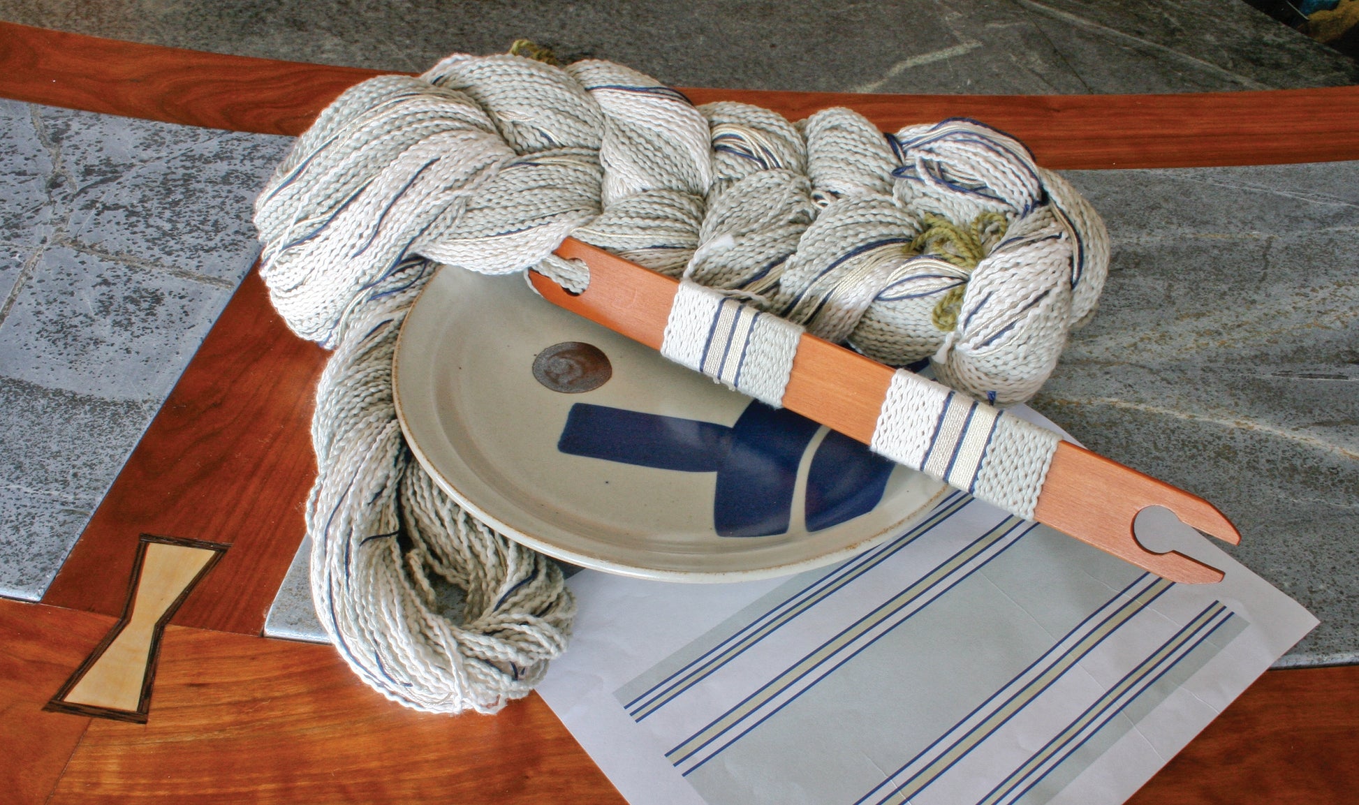 A coiled rope and a wooden weaving shuttle lie on top of an intricately crafted Rustic Mats, Runner & Napkins from Halcyon Yarn, featuring blue stripes and woven on a 4 shaft loom. This beautifully finished textile is displayed on a wooden table with stone inlay. A round ceramic plate with a cross design is also present, accompanied by elegant stoneware dishes.